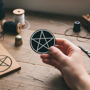 May include: A circular black patch with a white pentagram design, held by a hand. The image includes sewing equipment such as thread spools, a thimble, and scissors, suggesting a craft or DIY project. The patch is likely for clothing or accessories.