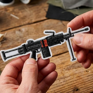 Può includere: Una toppa ricamata in bianco e nero a forma di mitragliatrice. La toppa presenta un design dettagliato con accenti rossi per le munizioni. La toppa è tenuta tra le mani su uno sfondo di legno.