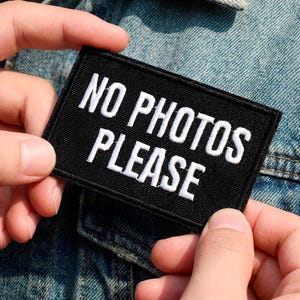 Może przedstawiać: Czarna, prostokątna naszywka z białym haftem "NO PHOTOS PLEASE". Naszywka jest prezentowana na kurtce dżinsowej.