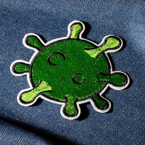 Op de afbeelding: Een groen geborduurde patch in de vorm van een virus, met een witte rand. De patch ligt op een blauwe denim achtergrond. Het virus heeft een rond lichaam met verschillende uitsteeksels.