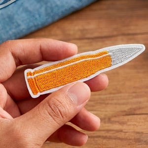 Op de afbeelding: Geborduurde patch in de vorm van een kogel. De patch heeft een witte rand, een zilveren punt en een oranje lichaam met een witte streep. De patch wordt in de hand van een persoon gehouden.