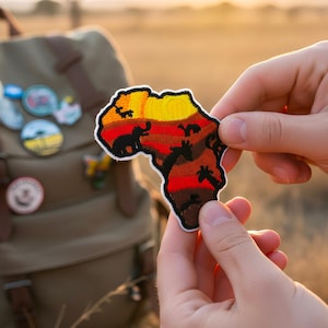 Può includere: Una toppa ricamata colorata a forma di continente africano, con un design a tramonto nei toni del giallo, arancione e rosso. La toppa è tenuta davanti a uno zaino con altre toppe attaccate.