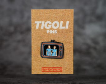 以下が含まれることがあります： Tigoli Pinsのエナメルピン。青いドレスを着た2人の人物が描かれたレトロなテレビです。ピンは茶色のカードにあり、「TIGOLI PINS」と「FOR ENTERTAINMENT PURPOSES ONLY」の文字が書かれています。