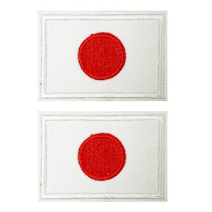 Puede incluir: Dos parches bordados con la bandera de Japón. Cada parche tiene un fondo rectangular blanco con un círculo rojo centrado, que representa el sol. Los parches tienen un borde cosido.
