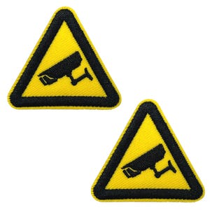 Könnte beinhalten: Zwei gelbe, dreieckige Aufnäher mit schwarzem Rand und einem schwarzen Überwachungskamera-Symbol. Die Aufnäher sind mit einem Warnsymbol bestickt, das auf Überwachung hinweist.