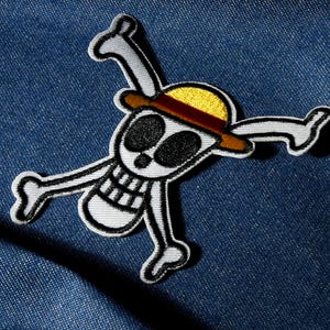 Op de afbeelding: Geborduurde patch met een witte schedel met zwarte oogkassen en een brede grijns, gekruist door twee botarmen. De schedel draagt een gele strohoed met een bruine band. De patch is zwart-wit omlijnd, op een blauwe denim achtergrond.