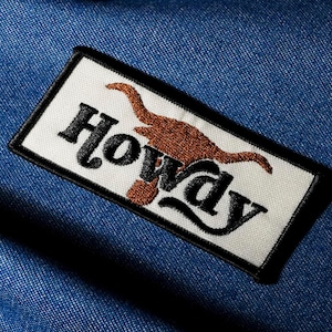 May include: Embroidered patch featuring the word "Howdy" in black script and a brown steer silhouette on a white background. The patch is edged in black, set against a blue denim fabric.