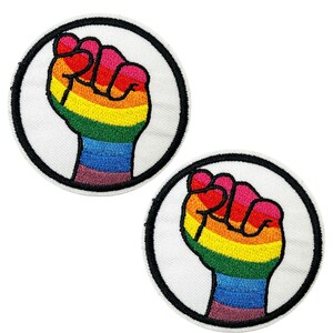 May include: Two embroidered patches showing a raised fist in rainbow colours. The fist is within a white circle with a black border. The patches have a sewn-on design.