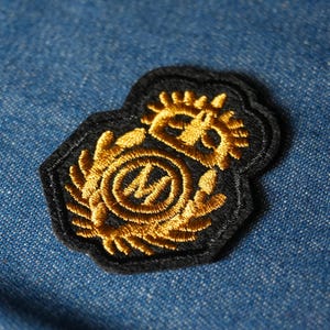 Peut inclure: Un écusson brodé noir avec un emblème doré représentant une lettre "M" stylisée dans un motif circulaire, entouré de détails ornés. L'écusson est posé sur un fond en denim bleu.