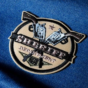 May include: Embroidered patch with the words "SHERIFF DEPARTMENT" in black on a tan circle. Two crossed pistols are above the text. A star and a green emblem are also present. The patch is on a blue denim background.