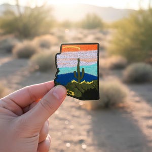 Könnte beinhalten: Gestickter Aufnäher in Form von Arizona, mit einer Wüstenlandschaft mit Kaktus, Bergen und einem farbenfrohen Sonnenuntergang. Der Aufnäher hat einen schwarzen Rand und wird vor einem verschwommenen Wüstenhintergrund gehalten.