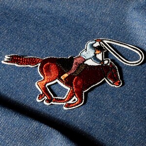 Puede incluir: Un parche bordado con un caballo y un jinete en tonos marrones, negros y azules. El parche tiene un borde blanco y un lazo en la parte superior. El caballo está en movimiento y el jinete lleva un sombrero.