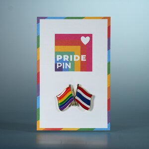 Op de afbeelding: Een Pride Pin-kaart met een regenboogrand en een kleurrijk vierkant met een wit hart en de woorden "PRIDE PIN". Twee vlagspelden zijn bevestigd: een regenboogvlag en een Thaise vlag.