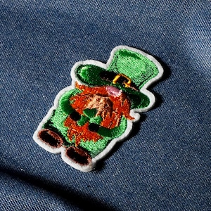 Könnte beinhalten: Gestickter Aufnäher mit einem Kobold in grünem Hut und Outfit, rotem Bart und braunen Schuhen. Der Aufnäher hat einen weißen Rand und ist auf einem blauen Jeanshintergrund platziert. Ein festlicher Artikel für St. Patrick's Day.