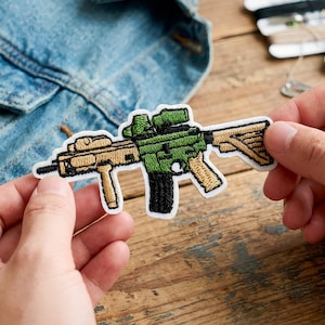 Può includere: Una toppa ricamata a forma di fucile, con colori verde, beige e nero. La toppa ha un bordo bianco ed è tenuta in mano. Lo sfondo comprende una giacca di jeans e una superficie di legno.