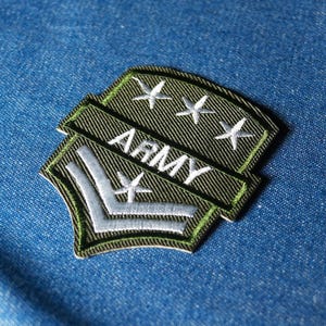 Op de afbeelding: Een geborduurd Army-patch met een groene achtergrond en witte tekst en sterren. De patch heeft het woord "ARMY" en een chevron-ontwerp, met drie sterren boven de tekst. De patch is geplaatst tegen een blauwe denim achtergrond.