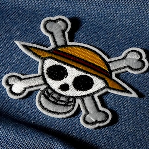 Op de afbeelding: Geborduurde patch met een schedel met een strohoed en gekruiste botten. De patch heeft een witte schedel met zwarte oogkassen en een lachende mond. De hoed is geel met een rode streep. De patch is omlijnd in zwart en grijs.