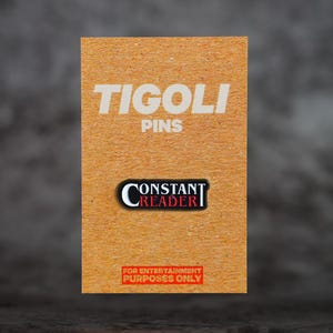Könnte beinhalten: Eine braune rechteckige Karte mit dem weißen Text "TIGOLI PINS". Darunter ein Anstecker mit "CONSTANT READER". Der untere Teil der Karte trägt den roten Text: "FOR ENTERTAINMENT PURPOSES ONLY".