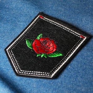 Könnte beinhalten: Schwarzer, gestickter Aufnäher mit einer roten Rose und grünen Blättern. Der Aufnäher hat einen weiß genähten Rand und rote Akzente an den Ecken. Auf blauem Denim-Hintergrund.
