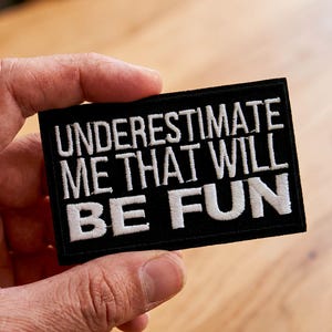 Könnte beinhalten: Schwarzer rechteckiger Aufnäher mit weiß gesticktem Text: „UNDERESTIMATE ME THAT WILL BE FUN“. Der Aufnäher wird von einer Hand gehalten.