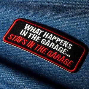 May include: Black rectangular embroidered patch with a red border and the text "WHAT HAPPENS IN THE GARAGE... STAYS IN THE GARAGE" in white and red lettering. The patch is on a blue denim background.