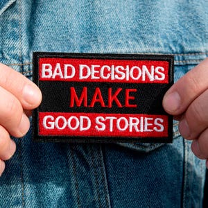 Könnte beinhalten: Ein rechteckiger, schwarz-roter Aufnäher mit dem weißen Schriftzug "BAD DECISIONS MAKE GOOD STORIES". Der Aufnäher wird vor einer Jeansjacke präsentiert.