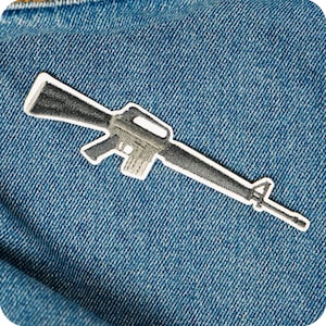 Könnte beinhalten: Ein gestickter Aufnäher mit einer detaillierten Darstellung eines Gewehrs. Der Aufnäher ist hauptsächlich grau und schwarz, mit weißer Umrandung. Er ist vor einem blauen Jeans-Hintergrund platziert.