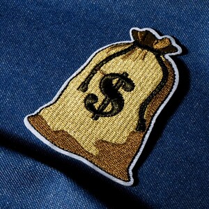 Pode incluir: Patch bordado com um desenho de saco de dinheiro. O saco é dourado com um símbolo de dólar preto no centro. O patch tem uma borda branca e está sobre um fundo de ganga azul.