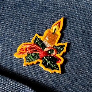 May include: Embroidered patch depicting a candle with a flame, holly leaves, and a red bow. The patch has a yellow border, set against a blue denim background. The design is detailed with green and red stitching.