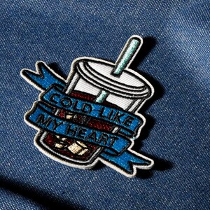 Peut inclure: Écusson brodé représentant une boisson froide dans un gobelet à emporter avec une paille. L'écusson comporte une bannière bleue avec les mots "COLD LIKE MY HEART" en lettres blanches. L'écusson est sur un fond en denim bleu.