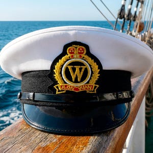 May include: A white naval cap with a black peak and band, adorned with a gold emblem displaying a 'W' and a crown. The cap sits on a wooden surface, with the sea visible in the background, evoking a maritime setting.