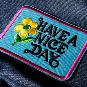Peut inclure: Écusson brodé avec un fond bleu clair et une bordure rose. L'écusson présente une fleur jaune avec des feuilles vertes et l'expression "HAVE A NICE DAY" en lettres noires. L'écusson est de forme rectangulaire.