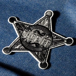 May include: Embroidered black and white sheriff badge patch with the word "WAR" in the center. The star-shaped patch has a white border and is set against a blue denim background.