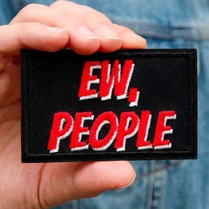 Może przedstawiać: Czarna prostokątna naszywka z napisem "EW, PEOPLE" wyszytym na czerwono z białymi obrysami. Naszywka jest trzymana w dłoni na tle dżinsowej kurtki.