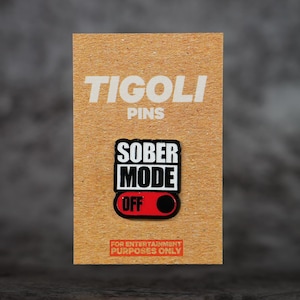 May include: A pin on a brown card with the text "TIGOLI PINS". The pin reads "SOBER MODE OFF" in black and white with a red toggle. The bottom of the card reads "FOR ENTERTAINMENT PURPOSES ONLY".