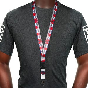 Op de afbeelding: Een lanyard met een herhalend patroon van rode, witte en blauwe rechthoeken. De lanyard wordt gedragen over een donkergrijs t-shirt. Het shirt heeft een wit logo op de linkermouw en een wit logo op de rechtermouw.