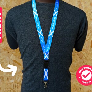 Op de afbeelding: Blauwe lanyard met het Schotse vlagontwerp en het woord "SCOTLAND" herhaald. Het heeft een zwarte plastic sluiting en een metalen clip. Het ontwerp is omkeerbaar.