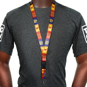 Puede incluir: Un cordón con un patrón repetitivo de cuadrados rojos, amarillos y azules. El cordón se lleva alrededor del cuello y cuelga por delante de una persona que lleva una camiseta gris oscuro. La camiseta tiene un logotipo blanco en cada manga.