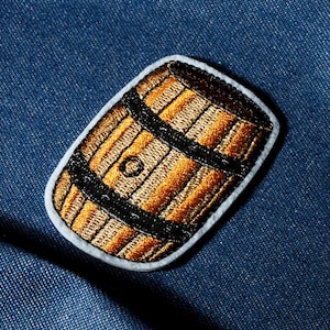 Puede incluir: Parche bordado con un diseño de barril marrón y dorado con bandas negras. El parche tiene un borde blanco y está sobre un fondo de mezclilla azul. El diseño del barril está detallado con líneas verticales.