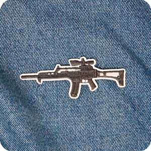 Könnte beinhalten: Ein schwarz-weiß gestickter Aufnäher eines Gewehrs mit Zielfernrohr, vor einem blauen Jeansstoff. Der Aufnäher hat einen weißen Rand, der eine klare Kontur bildet.