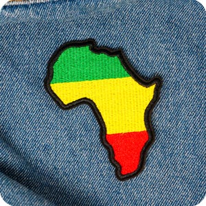 Op de afbeelding: Een geborduurde patch in de vorm van het Afrikaanse continent, met groene, gele en rode secties met een zwarte rand. De patch is geplaatst op een blauwe denim achtergrond.