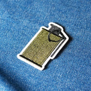 May include: Embroidered grenade-shaped patch. The patch has a green body with black and white details, and a white border. It is set against a blue denim background.