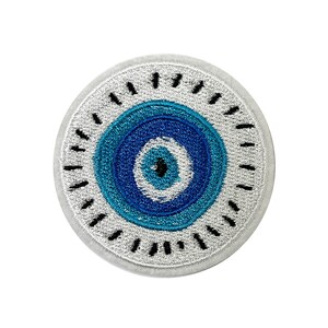 May include: Round embroidered patch with a blue and white evil eye design. The eye has concentric circles of blue, with a black pupil. The patch has a silver-grey border and black stitched details around the edge. Suitable for clothing or accessories.