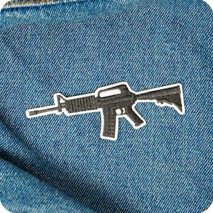 Puede incluir: Un parche negro y blanco con forma de rifle, con un borde blanco, está adherido a una tela vaquera azul. El parche detalla el contorno del rifle, incluyendo el cañón, el cargador y la culata.