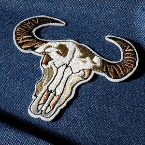 May include: Embroidered patch of a bull skull in white, brown, and gold. The patch has a detailed design with a white border, ideal for adding a unique touch to clothing or accessories.