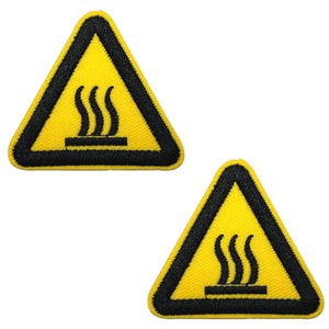 May include: Two embroidered patches in the shape of a yellow triangle with a black border. The patches feature a black symbol of rising steam above a horizontal line, indicating a hot surface. The patches are likely for clothing or accessories.