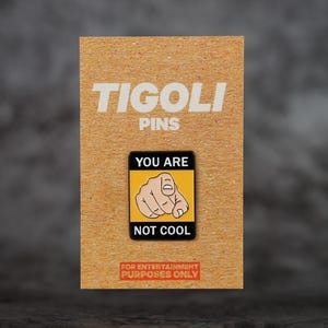 Può includere: Spilla Tigoli Pins su una scheda marrone. La spilla nera, gialla e bianca mostra una mano che indica lo spettatore e le parole «YOU ARE NOT COOL». La scheda recita «FOR ENTERTAINMENT PURPOSES ONLY».