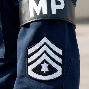 Könnte beinhalten: Nahaufnahme eines marineblauen Uniformärmels mit einem weiß-schwarzen Chevron-Abzeichen mit einem Stern. Über dem Ärmel befindet sich ein schwarzes Band mit den Buchstaben "MP" in Weiß.