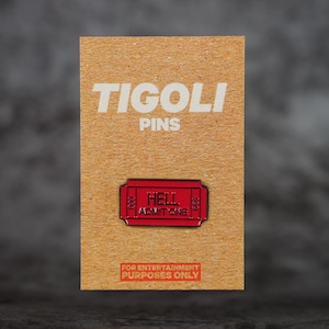 May include: A red enamel pin in the shape of a ticket that reads "HELL ADMIT ONE" on a brown card. The card has the text "TIGOLI PINS" at the top and "FOR ENTERTAINMENT PURPOSES ONLY" at the bottom.