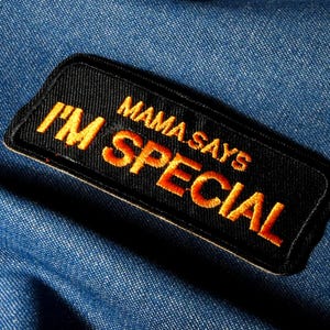 Op de afbeelding: Een zwart, rechthoekig embleem met de geborduurde tekst "MAMA SAYS I'M SPECIAL" in oranje en gele draad. Het embleem is geplaatst tegen een blauwe denim achtergrond, wat een contrast creëert tussen het donkere embleem en de lichtere stof.